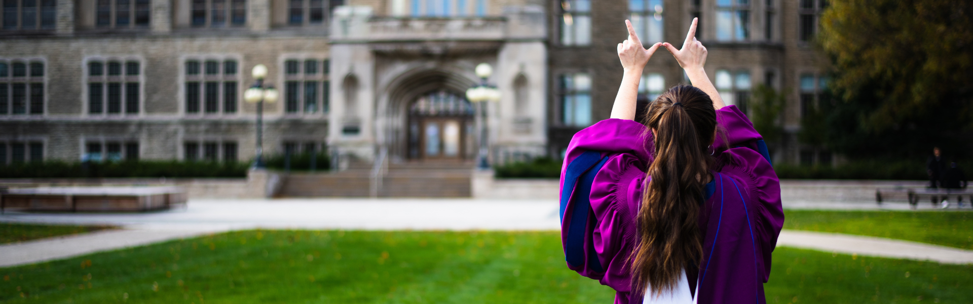 A graduate standing on campus in regalia creating a "W" with their hands