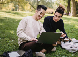 Two students sitting on the grass with their laptops and notebooks